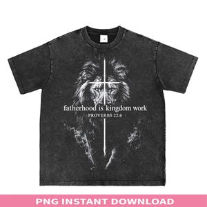 May include: A black acid-wash t-shirt featuring a white graphic of a lion's face overlaid with a cross. The text reads, "fatherhood is kingdom work" and "PROVERBS 22:6" in white lettering centered on the chest of the garment.