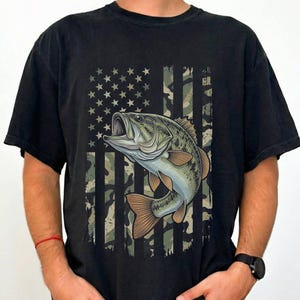 May include: Black t-shirt featuring a graphic of a largemouth bass with an open mouth, set against a camouflage and star-spangled flag design. The fish is detailed with shades of green, brown, and blue. The shirt is a casual wear item.