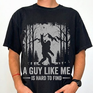 May include: Black t-shirt featuring a Bigfoot graphic in a forest setting, with the text "A Guy Like Me Is Hard To Find". The design includes a silhouette of Bigfoot with a raised hand gesture, trees, and mountains.