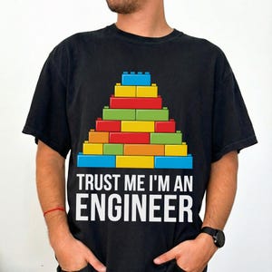 May include: Black t-shirt featuring a colorful block design in the shape of a pyramid. The text "TRUST ME I'M AN ENGINEER" is printed in white below the design. The shirt is a casual wear item.