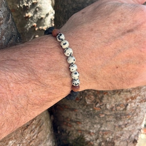 May include: A beaded bracelet featuring a sequence of round white beads with black spots, interspersed with two smooth brown wooden beads and two textured black porous beads, arranged in a single strand on an elastic cord.