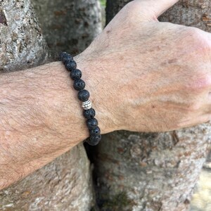 May include: A beaded bracelet featuring round, porous black lava stone beads strung together. A single, cylindrical silver-toned metal bead with a textured, grooved surface is positioned among the black beads, providing a metallic contrast.