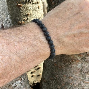 May include: A stretch bracelet composed of small, round, porous black lava stone beads. The beads feature a rough, textured surface and are strung together in a single continuous loop, creating a dark, uniform appearance around the wrist.