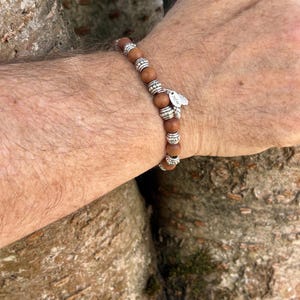 May include: A beaded bracelet featuring smooth, round brown wooden beads alternating with textured, silver-toned metal spacer beads. A small, oval-shaped silver-toned metal charm with engraved text is attached to the side of the bracelet.