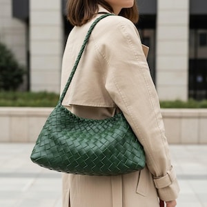 Pode incluir: Uma bolsa de ombro verde em couro tran&ccedil;ado com uma al&ccedil;a tran&ccedil;ada. A bolsa &eacute; de cor verde esmeralda profunda e tem formato retangular com cantos arredondados. A bolsa &eacute; usada sobre um trench coat bege.