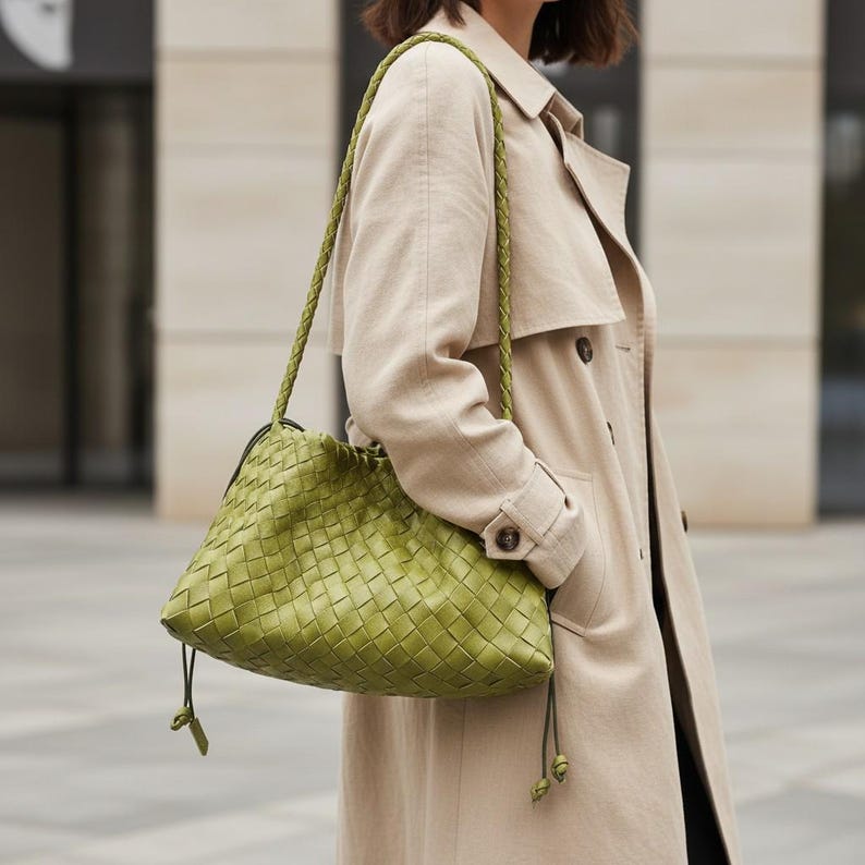 Pode incluir: Uma bolsa de couro verde tecido com al&ccedil;as de ombro tran&ccedil;adas. A bolsa tem um fecho de cord&atilde;o e &eacute; usada no ombro. A bolsa &eacute; de cor verde brilhante e tem uma apar&ecirc;ncia texturizada. A pessoa na imagem est&aacute; usando um trench coat bege.