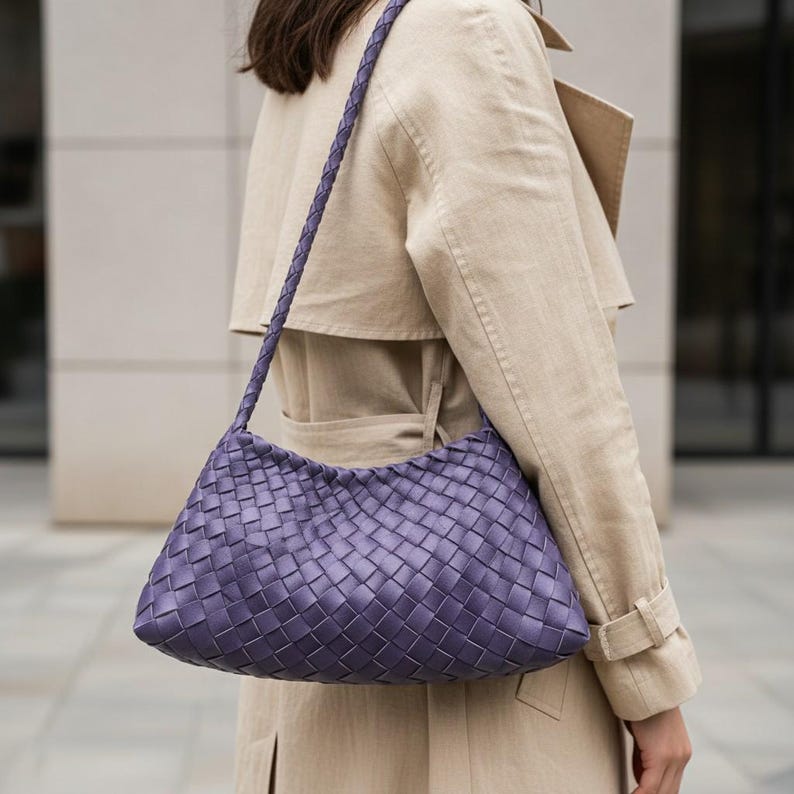 Pode incluir: Uma bolsa de ombro tecida, cor lavanda, com uma al&ccedil;a tran&ccedil;ada. A bolsa tem formato retangular com bordas arredondadas. A bolsa &eacute; usada no ombro. O fundo apresenta um trench coat bege.