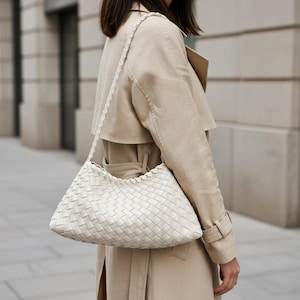 Pode incluir: Bolsa de ombro em couro tran&ccedil;ado branco sujo com uma al&ccedil;a tran&ccedil;ada. A pessoa est&aacute; usando um trench coat bege. O padr&atilde;o tran&ccedil;ado da bolsa &eacute; vis&iacute;vel, e o estilo geral &eacute; minimalista e chique.
