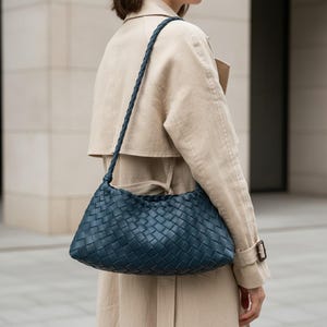 Pode incluir: Uma bolsa de ombro de couro tran&ccedil;ado azul-petr&oacute;leo com uma al&ccedil;a tran&ccedil;ada. A bolsa tem formato retangular com bordas arredondadas e um formato macio e folgado. A bolsa &eacute; usada sobre um trench coat bege.