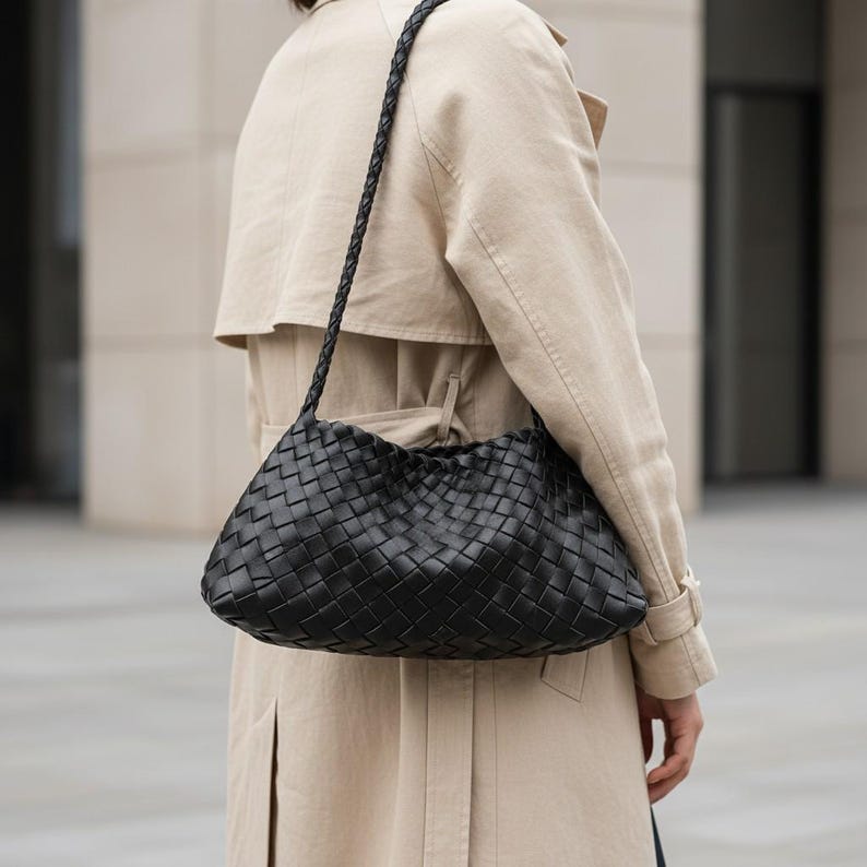 Pode incluir: Bolsa de ombro preta em couro tran&ccedil;ado com al&ccedil;a tran&ccedil;ada. A bolsa tem um estilo macio e folgado e &eacute; usada no ombro. A pessoa est&aacute; usando um trench coat bege.