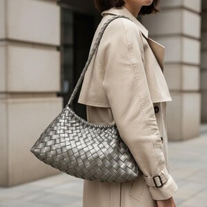 Pode incluir: Uma bolsa de ombro prateada tecida com uma al&ccedil;a tran&ccedil;ada. A bolsa tem formato retangular com bordas arredondadas. A bolsa &eacute; feita de material met&aacute;lico e &eacute; usada no ombro. O fundo mostra um trench coat bege.