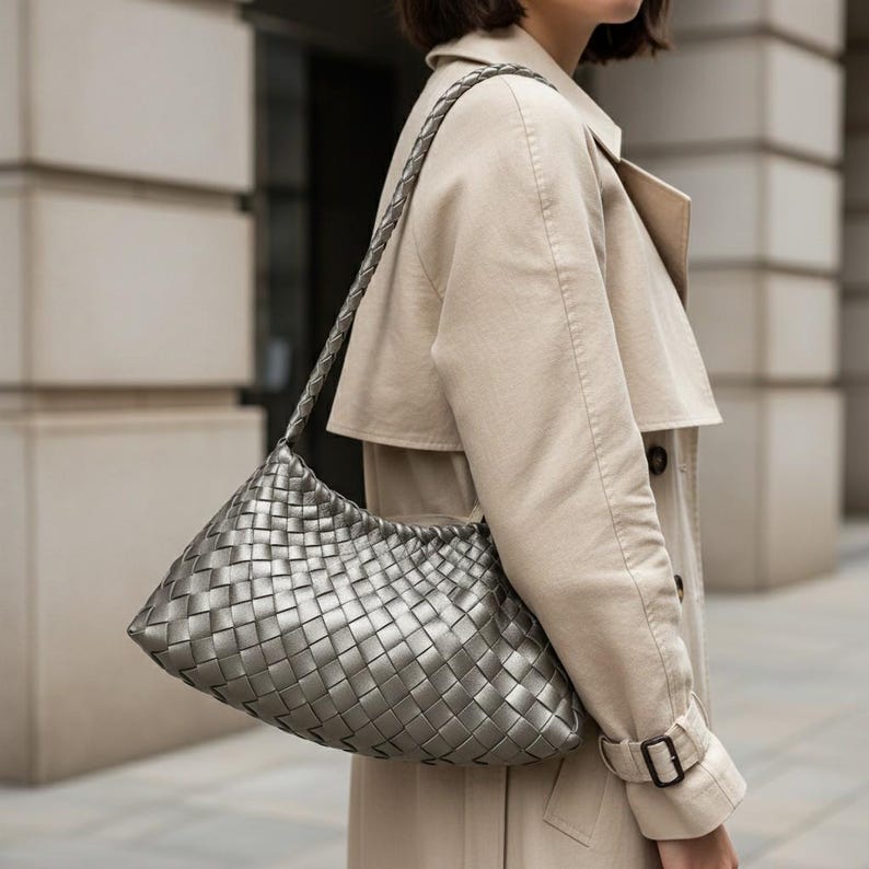 Pode incluir: Uma bolsa de ombro prateada tecida com uma al&ccedil;a tran&ccedil;ada. A bolsa tem formato retangular com bordas arredondadas. A bolsa &eacute; feita de material met&aacute;lico e &eacute; usada no ombro. O fundo mostra um trench coat bege.