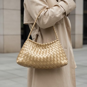 Pode incluir: Uma bolsa dourada tecida com uma al&ccedil;a tran&ccedil;ada. A bolsa tem formato retangular com cantos arredondados. A bolsa &eacute; segurada por uma pessoa que usa um trench coat bege. O padr&atilde;o tecido da bolsa &eacute; vis&iacute;vel.