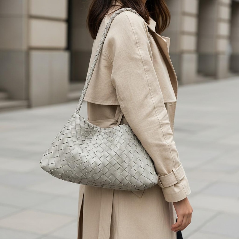 Pode incluir: Uma bolsa de ombro de couro tran&ccedil;ado cinza claro com uma al&ccedil;a tran&ccedil;ada. A bolsa tem formato arredondado e &eacute; usada no ombro. A bolsa &eacute; combinada com um trench coat bege.
