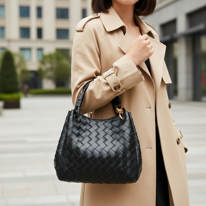 Peut inclure: Un sac &agrave; main noir dot&eacute; d'un ext&eacute;rieur en cuir tress&eacute;, d'une base structur&eacute;e et d'une anse unique avec des d&eacute;tails tress&eacute;s. Le sac comprend des attaches en m&eacute;tal dor&eacute; reliant la poign&eacute;e au corps principal de l'accessoire de mode.