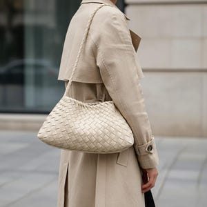 Pode incluir: Bolsa de m&atilde;o de couro tran&ccedil;ado creme com uma al&ccedil;a tran&ccedil;ada. A bolsa tem formato retangular com bordas arredondadas e um padr&atilde;o tran&ccedil;ado. A pessoa est&aacute; usando um trench coat bege.