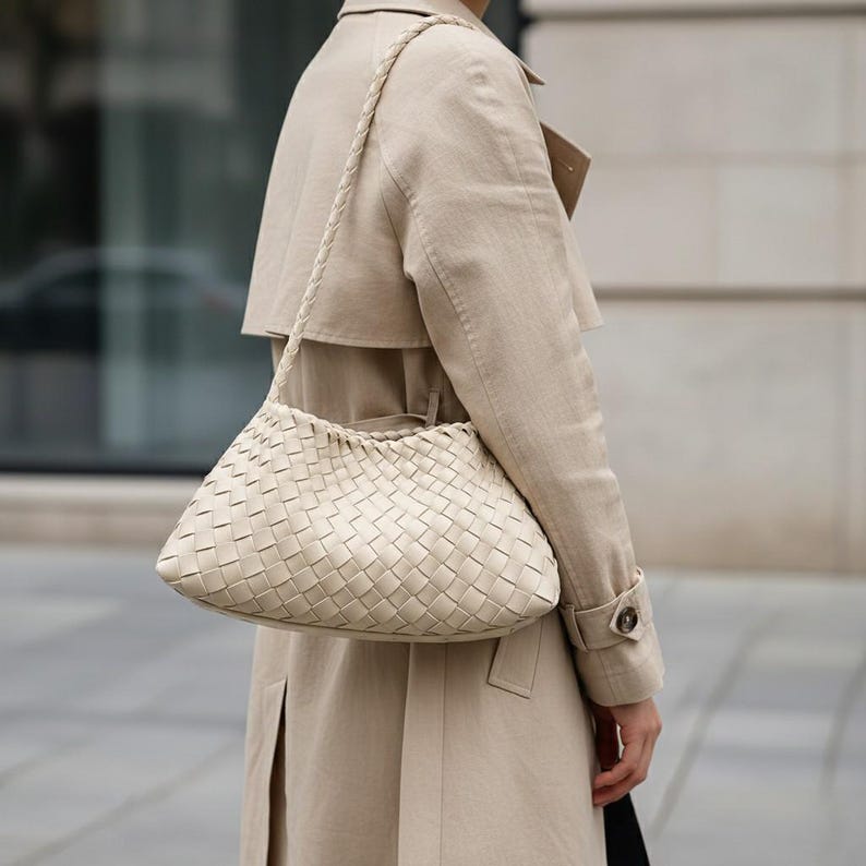 Pode incluir: Bolsa de m&atilde;o de couro tran&ccedil;ado creme com uma al&ccedil;a tran&ccedil;ada. A bolsa tem formato retangular com bordas arredondadas e um padr&atilde;o tran&ccedil;ado. A pessoa est&aacute; usando um trench coat bege.