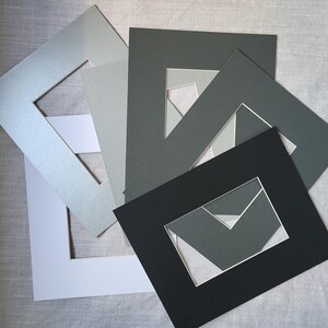 May include: A collection of rectangular paper picture mounts in various shades of grey, black, and white. Each mount features a centred rectangular aperture with a bevelled edge, layered in a fan-like arrangement to display the different colour options.