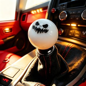 May include: A white shift knob with a black, grinning skull face design. The knob is round and sits atop a black, textured shift boot with red stitching. The interior of the car is red and black.