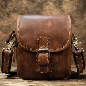 May include: A brown leather crossbody bag with a flap closure and adjustable strap. The bag features a metal buckle and stitching details. The strap has a brown and dark brown pattern. The bag is designed to be worn over the shoulder.