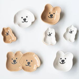 May include: A collection of ceramic dog-shaped dishes in various sizes and colors. The dishes are white and tan, with black facial features. The dishes are arranged on a white surface. These dishes are ideal for serving snacks or as decorative items.