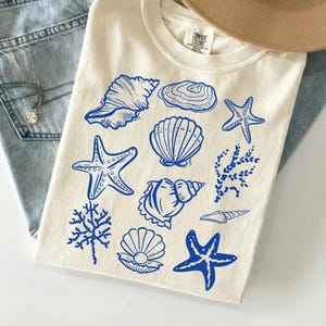 Könnte beinhalten: Cremefarbenes T-Shirt mit blauen Illustrationen von Muscheln, Seesternen und Korallen. Das T-Shirt ist auf einer hellblauen Jeans gefaltet, mit einem beigen Hut im Hintergrund. Das T-Shirt hat ein Comfort Colors-Etikett.