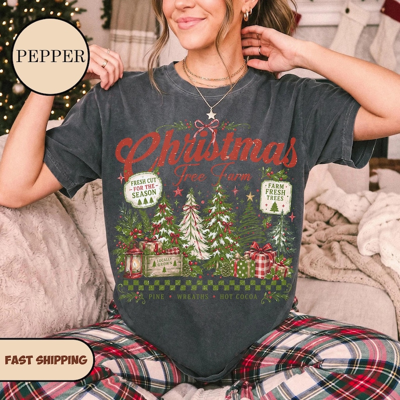 May include: A charcoal gray short-sleeve t-shirt featuring a graphic print with the text Christmas Free Farm. The design includes illustrations of pine trees, wrapped gifts, a lantern, and signs reading Fresh Cut For The Season and Farm Fresh Trees.