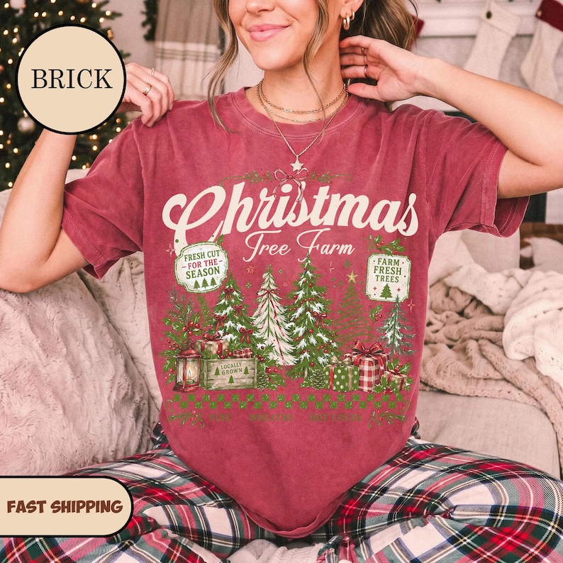 May include: A brick red short-sleeve t-shirt featuring a graphic print with the text Christmas Tree Farm, Fresh Cut For The Season, and Farm Fresh Trees. The design includes illustrations of evergreen trees, gift boxes, and a lantern.