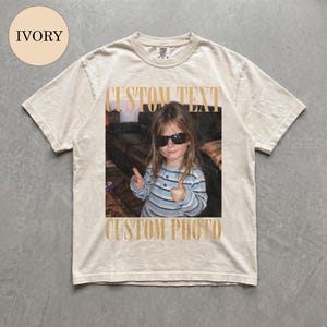 May include: An ivory short-sleeve t-shirt featuring a central graphic print of a child wearing sunglasses and a blue striped shirt, making a double thumbs-up gesture. The text CUSTOM TEXT appears above the photo and CUSTOM PHOTO below it.