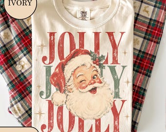 May include: An ivory t-shirt featuring a graphic of a winking Santa Claus wearing a red hat with holly berries. The text JOLLY JOLLY JOLLY is printed in a distressed red and green font, accented by small star shapes across the front of the shirt.