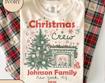 May include: An ivory t-shirt featuring a graphic of a Christmas tree and a fireplace. The text reads Christmas Crew, Johnson Family, New York, NY, and 2026. The design includes red and green ornaments, snowflakes, and gifts in a festive style.