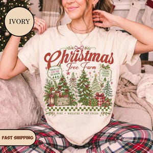 May include: An ivory t-shirt featuring a graphic with the text "Christmas Tree Farm" in red script. The design includes green pine trees, wrapped gifts, a lantern, and text reading "Fresh Cut For The Season," "Farm Fresh Trees," and "Pine Wreaths Hot Cocoa."