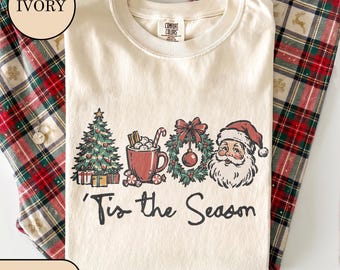 May include: An ivory t-shirt featuring a graphic print of a Christmas tree, a mug of hot cocoa, a wreath, and Santa Claus. Below the illustrations, the text reads 'Tis the Season in a black script font. The shirt has a crew neckline and short sleeves.