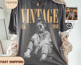 May include: A charcoal gray short-sleeve t-shirt featuring a vintage-style graphic of a person wearing sunglasses and sitting on a skateboard. The text reads VINTAGE EST. 1980 in large tan letters. A small inset photo shows the original reference image.