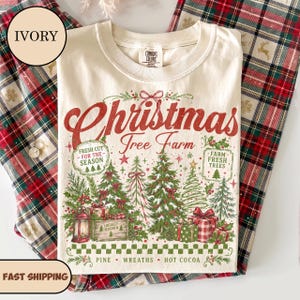 May include: An ivory t-shirt featuring a red and green graphic with the text Christmas Tree Farm, Fresh Cut For The Season, Farm Fresh Trees, and Locally Grown. The design includes illustrations of pine trees, wrapped gifts, and a lantern.