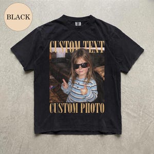 May include: A black t-shirt featuring a graphic print of a child wearing sunglasses and a blue striped shirt, making a double thumbs-up gesture. The text CUSTOM TEXT appears above the image and CUSTOM PHOTO appears below in a tan serif font.