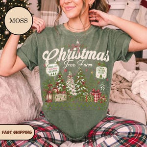 May include: A moss green short-sleeve t-shirt featuring a graphic print with the text Christmas Tree Farm, Fresh Cut For The Season, and Farm Fresh Trees. The design includes illustrations of pine trees, gift boxes, and a lantern.