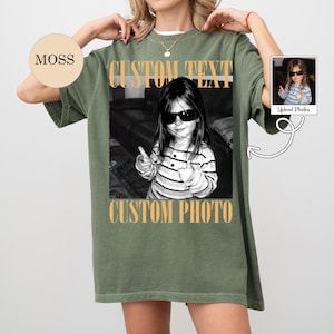 May include: An olive green oversized t-shirt featuring a black and white photograph of a child wearing sunglasses and a striped shirt. The text CUSTOM TEXT appears above the photo and CUSTOM PHOTO appears below it in a bold, tan, serif font.