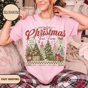May include: A light pink short-sleeve t-shirt featuring a graphic print with the text "Christmas Tree Farm" in red script. The design includes green pine trees, gift boxes, a lantern, and two rectangular signs reading "Fresh Cut For The Season" and "Farm Fresh Trees".