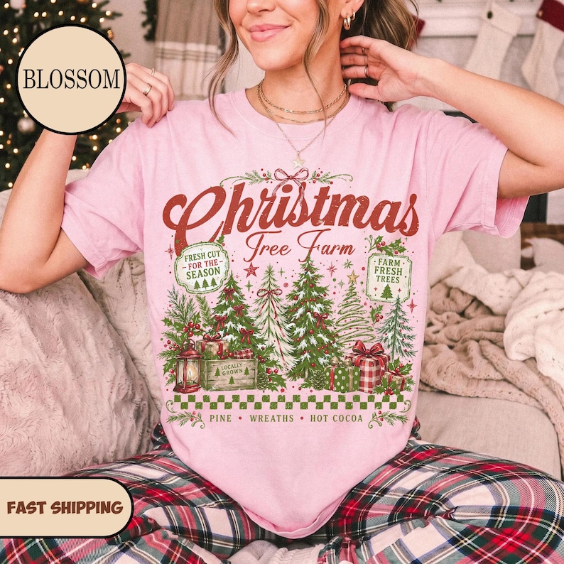 May include: A light pink short-sleeve t-shirt featuring a graphic print with the text "Christmas Tree Farm" in red script. The design includes green pine trees, gift boxes, a lantern, and two rectangular signs reading "Fresh Cut For The Season" and "Farm Fresh Trees".