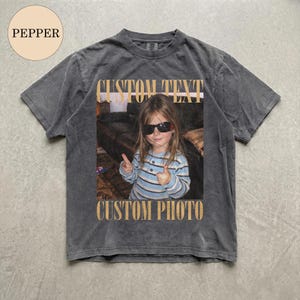 May include: A charcoal grey short-sleeve t-shirt featuring a central rectangular graphic of a child wearing sunglasses and a blue striped shirt, making a peace sign. The text CUSTOM TEXT appears above the image and CUSTOM PHOTO below it.