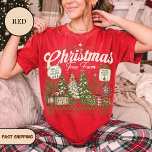 May include: A red short-sleeve t-shirt featuring a graphic print with the text Christmas Tree Farm, Fresh Cut For The Season, and Farm Fresh Trees. The design includes illustrations of pine trees, gift boxes, and a lantern in green and red.