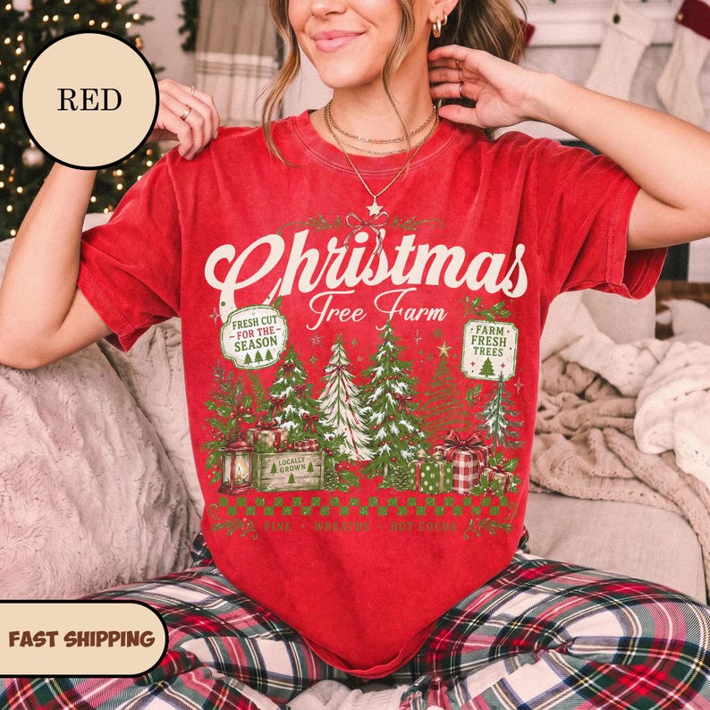 May include: A red short-sleeve t-shirt featuring a graphic print with the text Christmas Tree Farm, Fresh Cut For The Season, and Farm Fresh Trees. The design includes illustrations of pine trees, gift boxes, and a lantern in green and red.
