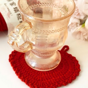 Puede incluir: Una taza de cristal iridiscente transparente con una superficie facetada y texturizada y un asa decorativa, apoyada sobre un posavasos redondo de ganchillo rojo brillante con un pequeño lazo en el borde superior. La taza tiene un relieve.