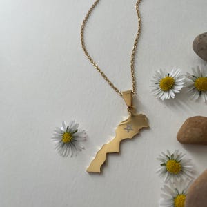 Peut inclure: Un collier doré avec un pendentif en forme de carte du Maroc. Le pendentif présente un motif d'étoile. Le collier est présenté avec des marguerites blanches et de petites pierres sur une surface blanche.