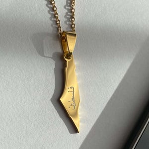 Könnte beinhalten: Goldfarbener Anhänger in Form von Palästina mit dem arabischen Wort „Palästina“. Der Anhänger hängt an einer zarten Goldkette. Das Bild ist vor einem neutralen Hintergrund platziert, der den Schmuck hervorhebt.