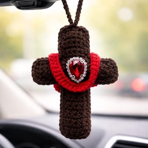 May include: A brown crocheted cross car charm with a red ribbon and a large, red jewel in the center. The charm is suspended by a brown cord, and is hanging in front of a blurred background.