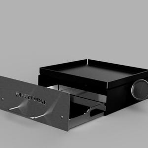 Puede incluir: Una parrilla de metal negro con un cajón extraíble. La parrilla tiene una parte superior plana y un dial circular en el lateral. El cajón tiene el texto "VULCAN FORGE" grabado. La parrilla está sobre un fondo gris.