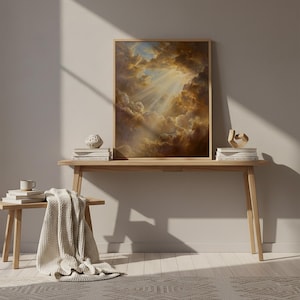 May include: A framed artwork depicting a dramatic sky with golden light piercing through clouds. The artwork is displayed on a light wood console table, alongside decorative objects and books. A neutral-toned throw blanket and a coffee cup add to the cozy aesthetic.