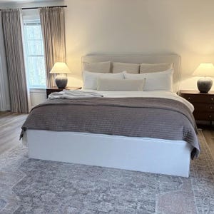 May include: A bedroom featuring a bed with a white upholstered frame, a dark gray textured quilt, and multiple white pillows. Two dark wood nightstands hold lamps with white shades. Long beige curtains hang by a window.